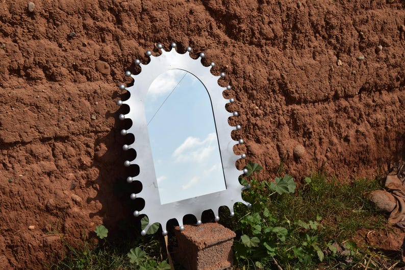 Op de afbeelding: Een decoratieve spiegel met een uniek, tandwielachtig frame. Het zilverkleurige frame heeft een reeks afgeronde tanden en kleine, zilverkleurige bollen. De spiegel reflecteert een blauwe lucht met witte wolken. De spiegel wordt buiten tentoongesteld.