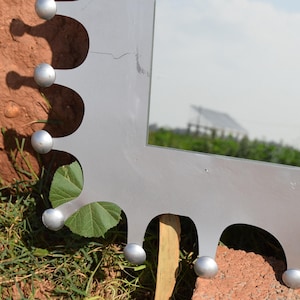 Op de afbeelding: Een zilverkleurige spiegel met een uniek geschulpte rand. De spiegel heeft kleine, ronde, zilveren accenten langs de rand. De achtergrond omvat groen gras, bladeren en een gebouw in de verte.