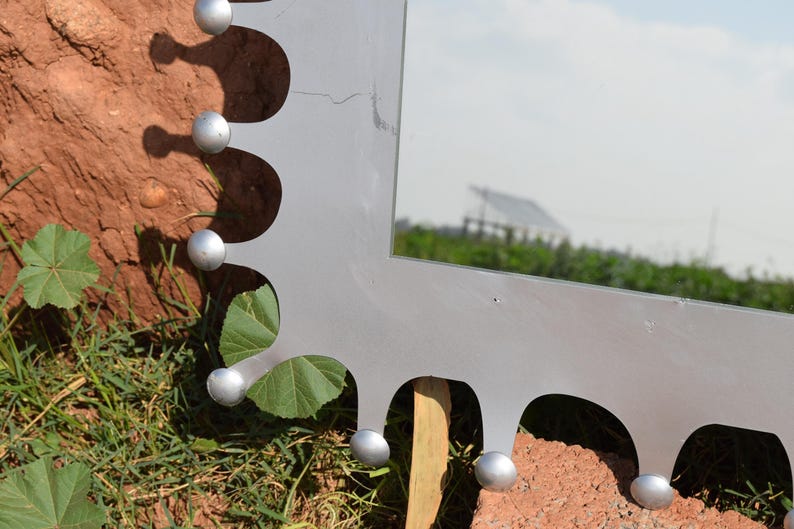 Op de afbeelding: Een zilverkleurige spiegel met een uniek geschulpte rand. De spiegel heeft kleine, ronde, zilveren accenten langs de rand. De achtergrond omvat groen gras, bladeren en een gebouw in de verte.
