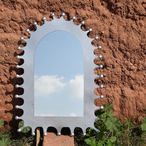 Op de afbeelding: Een zilverkleurige boogspiegel met een decoratieve rand voorzien van kleine, gelijkmatig verdeelde bolvormige uitsteeksels langs de gehele buitenrand. Het reflecterende oppervlak toont een blauwe lucht met witte wolken.