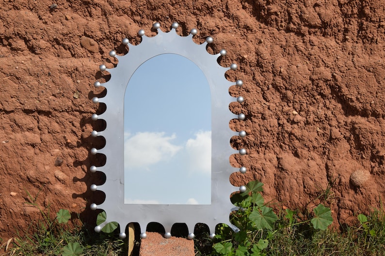 Op de afbeelding: Een zilverkleurige boogspiegel met een decoratieve rand voorzien van kleine, gelijkmatig verdeelde bolvormige uitsteeksels langs de gehele buitenrand. Het reflecterende oppervlak toont een blauwe lucht met witte wolken.