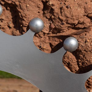 Op de afbeelding: Close-up van een decoratieve metalen kroon met vier zilveren bollen. De kroon is lichtgrijs van kleur en heeft een gekartelde rand. De achtergrond is een getextureerd, roodbruin oppervlak.