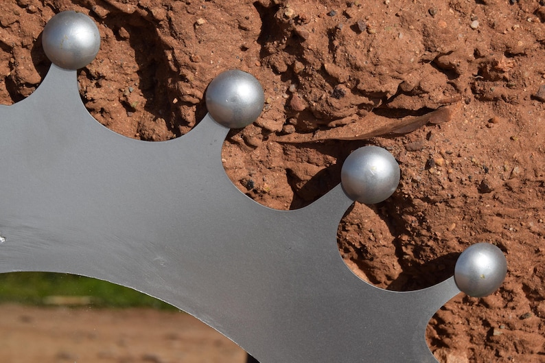 Op de afbeelding: Close-up van een decoratieve metalen kroon met vier zilveren bollen. De kroon is lichtgrijs van kleur en heeft een gekartelde rand. De achtergrond is een getextureerd, roodbruin oppervlak.