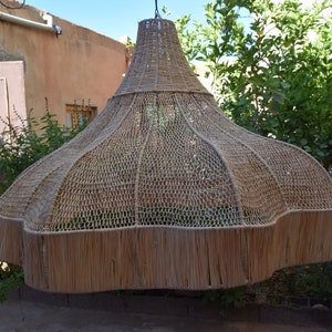 May include: A large, woven, light brown lampshade hangs from the ceiling in a courtyard. The lampshade is made of natural materials and has a fringed edge.