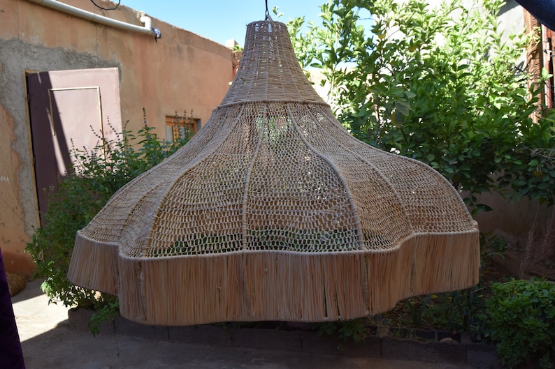 May include: A large, woven, light brown lampshade hangs from the ceiling in a courtyard. The lampshade is made of natural materials and has a fringed edge.