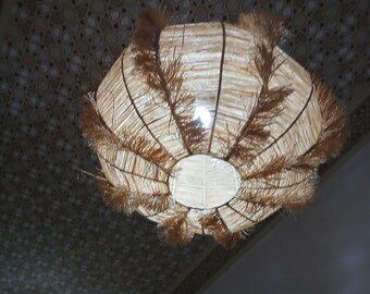 May include: A woven pendant light fixture with a natural, beige color. The light has a round shape with a decorative fringe around the edges. The light is hanging from a ceiling with a patterned design.