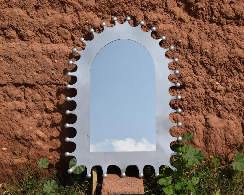 Op de afbeelding: Een decoratieve zilverkleurige spiegel met een gewelfde bovenkant en een uniek, geschulpte randontwerp. Kleine, ronde zilveren accenten sieren de rand. De spiegel reflecteert een blauwe lucht met wolken. De spiegel is gemonteerd op een roodbruine getextureerde muur.