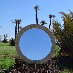 Op de afbeelding: Een ronde spiegel met een geweven, lichtgekleurde lijst staat in een veld met palmbomen onder een heldere blauwe lucht. De spiegel reflecteert de lucht, wat een opvallend contrast creëert met de natuurlijke omgeving.