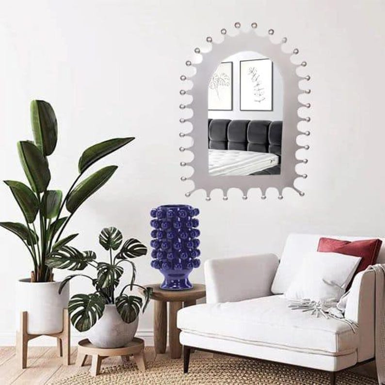 Op de afbeelding: Een wandspiegel met een geschulpt, kroonachtig frame met kleine bolvormige accenten langs de rand. De spiegel reflecteert een slaapkamerinterieur met een donkergrijs hoofdeinde en twee ingelijste lijntekeningen aan de muur.