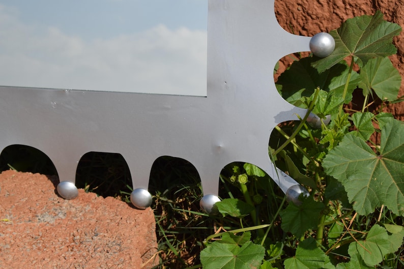 Op de afbeelding: Een grote, zilveren metalen sculptuur met een zaagtandrand, versierd met kleine, zilveren bollen. De sculptuur staat tegen een achtergrond van groen gebladerte en een roodbruin bakstenen oppervlak, met een bewolkte lucht op de achtergrond.
