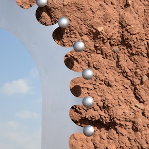 Op de afbeelding: Een close-up van een metalen sculptuur met een halfronde vorm en een reeks gelijkmatig verdeelde, bolvormige zilveren accenten. De sculptuur staat tegen een textuurrijke, roodbruine achtergrond, met een glimp van een blauwe lucht.