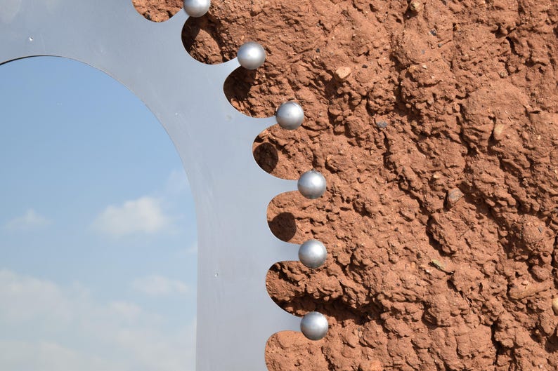 Op de afbeelding: Een close-up van een metalen sculptuur met een halfronde vorm en een reeks gelijkmatig verdeelde, bolvormige zilveren accenten. De sculptuur staat tegen een textuurrijke, roodbruine achtergrond, met een glimp van een blauwe lucht.