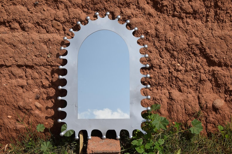 Op de afbeelding: Een decoratieve zilverkleurige spiegel met een uniek geschulpte randontwerp. De spiegel heeft een rechthoekige vorm met een gewelfde bovenkant en is gemonteerd op een bakstenen en aarden muur. De lucht wordt weerspiegeld in de spiegel.
