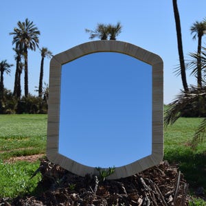 Op de afbeelding: Een rechthoekige spiegel met een afgeronde bovenkant, omlijst in een lichtbeige geweven materiaal. De spiegel reflecteert een heldere blauwe lucht. De spiegel staat tegen een achtergrond van groen gras en palmbomen.