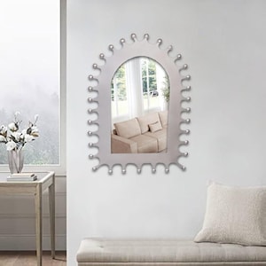 Op de afbeelding: Een boogvormige wandspiegel met een zilveren frame voorzien van een reeks kleine, gelijkmatig verdeelde bolvormige uitsteeksels langs de gehele buitenrand. Het spiegeloppervlak toont een beige bank en een raam.