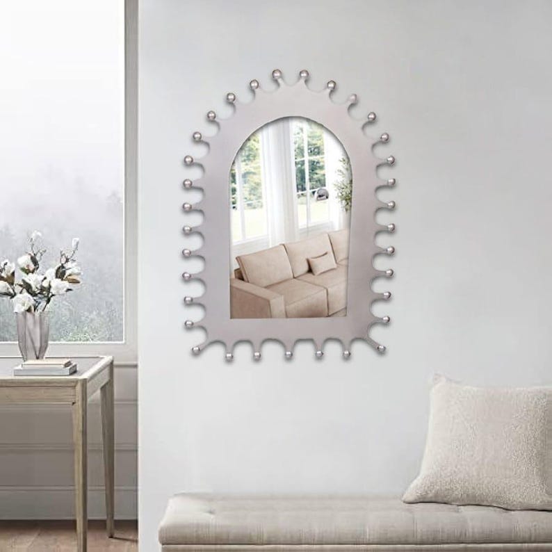 Op de afbeelding: Een boogvormige wandspiegel met een zilveren frame voorzien van een reeks kleine, gelijkmatig verdeelde bolvormige uitsteeksels langs de gehele buitenrand. Het spiegeloppervlak toont een beige bank en een raam.