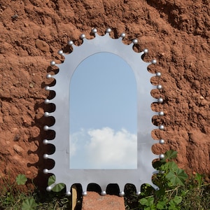 Op de afbeelding: Een zilverkleurige, boogvormige spiegel met een uniek, tandwielachtig frame. Het frame is versierd met kleine, ronde accenten. De spiegel reflecteert een blauwe lucht met wolken, tegen een getextureerde, roodbruine muur.