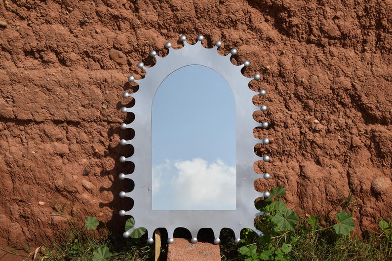 Op de afbeelding: Een zilverkleurige, boogvormige spiegel met een uniek, tandwielachtig frame. Het frame is versierd met kleine, ronde accenten. De spiegel reflecteert een blauwe lucht met wolken, tegen een getextureerde, roodbruine muur.