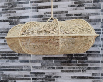 May include: A beige woven pendant light, oval in shape with a netted design. It hangs from a thin, beige cord. The background is a grey and white brick wall. The light fixture is suitable for a living room.