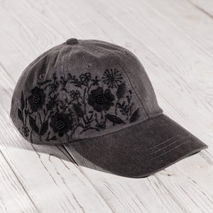 May include: A gray baseball cap with black embroidered floral designs. The cap has a curved brim and a black stitching detail.