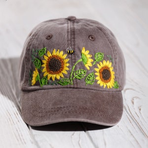 Op de afbeelding: Een bruine baseballpet met geborduurde zonnebloemen en een bij. De pet heeft een gebogen klep en een mesh achterkant.