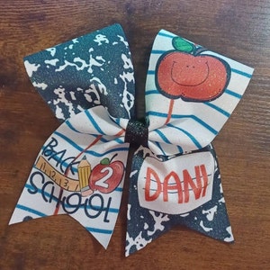 Może przedstawiać: Duża, błyszcząca kokarda do włosów o tematyce szkolnej. Kokarda ma wzór papieru w kratkę, grafikę jabłka oraz tekst "BACK 2 SCHOOL" i "DANI". Kokarda jest biała, niebieska, czerwona i czarna.