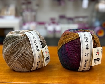 May include: Two round balls of Noro yarn with paper labels. The left ball features variegated earth tones of brown, beige, and grey. The right ball displays a vibrant mix of deep purple, magenta, and mustard yellow. Both labels include Japanese text.