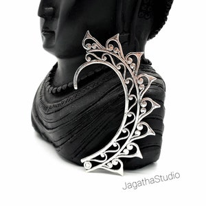 May include: Silver ear cuff with a delicate, intricate design. The cuff is shaped like a crescent moon and features a floral pattern.