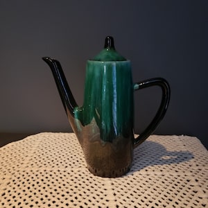 Puede incluir: Una tetera de cerámica verde y negra con un pico y un asa. La tetera tiene un esmalte que parece agua corriente.
