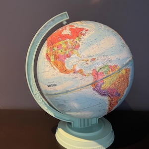 Może przedstawiać: Jasnoniebieski globus z serii World Scholar z szczegółową mapą świata. Globus jest zamontowany na pasującej jasnoniebieskiej podstawie i ma pierścień południka. Klasyczne narzędzie edukacyjne.