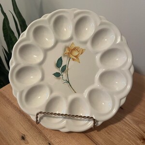 Pode incluir: Um prato de ovos de cerâmica branca com 12 reentrâncias individuais para servir ovos recheados. O prato apresenta uma rosa amarela com folhas verdes no centro.