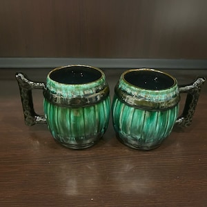 May include: Two ceramic beer steins with a green and black barrel design. The mugs have black handles and a gold rim. The interior of the mugs is black. The steins are on a dark brown surface.