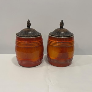 May include: Two ceramic jars with lids. The jars are a gradient of orange and brown with horizontal ridges. The lids are dark brown with a textured, cone-shaped finial. The jars are on a white surface.