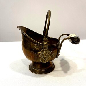May include: A brass coal scuttle with a pedestal base, a curved handle, and a rear handle featuring a white ceramic grip. The brass body has a polished finish and decorative metal plates at the base of the main handle, showing a relief design.