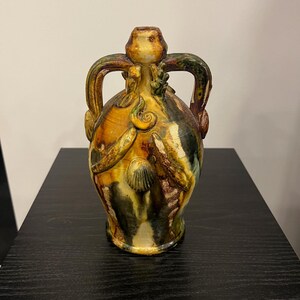 Peut inclure: Un vase en céramique vernissée doté d'un col étroit, de deux anses arquées et d'un corps bombé. La surface présente une palette de couleurs marbrées ambre, vert foncé et brun, rehaussée de reliefs décoratifs incluant un motif de coquillage.