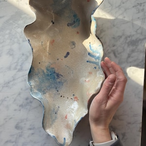 May include: A handmade ceramic serving dish with an irregular, organic shape. The dish features a speckled, off-white base with blue and red paint splatters. The rim is outlined in blue.