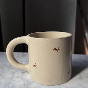 Może przedstawiać: Beżowy ceramiczny kubek z zaokrąglonym uchwytem. Kubek jest ozdobiony małymi brązowymi ilustracjami królików. Kubek stoi na marmurowej powierzchni.