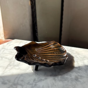 Può includere: Un piatto in ceramica a forma di conchiglia con finitura lucida. La superficie presenta una sfumatura dal marrone scuro al nero con creste prominenti che si irradiano dalla base. Il piatto è sostenuto da piccoli piedini.