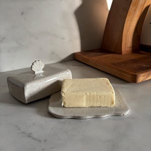 Op de afbeelding: Een rechthoekige botervloot van keramiek met een gespikkeld gebroken wit glazuur en een kleine schelpvormige handgreep op het deksel. De botervloot bestaat uit een platte bodem en een afneembaar deksel, getoond met een blok gele boter.