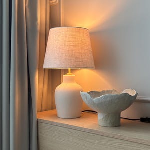May include: A table lamp with a textured off-white ceramic base and a tapered fabric lampshade emitting a warm glow, positioned next to a wide, shallow, off-white ceramic bowl with a rough, uneven, organic-shaped rim and a pedestal base.