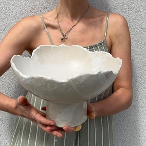 May include: A large, off-white ceramic bowl with a pedestal base. The bowl has a scalloped rim and a textured surface. The bowl is held by a person wearing a green and white striped dress.