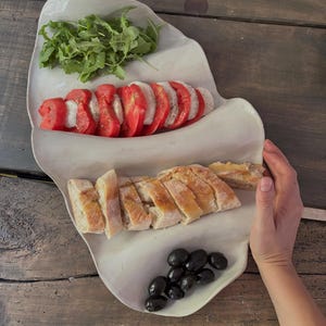 Pode incluir: Uma travessa de servir em cerâmica de cor branco-sujo com formato orgânico e duas secções distintas. A secção superior contém tomates vermelhos fatiados, queijo mozzarella branco e rúcula verde. A secção inferior contém pão baguette fatiado e azeitonas pretas.