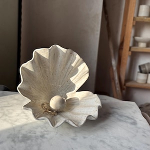 May include: A decorative, open clam shell-shaped bowl made of a light-coloured stone. Inside the shell is a small, round, speckled stone and a gold-coloured ring. The bowl sits on a white and grey marble surface.