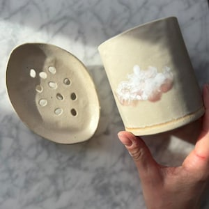 May include: A beige ceramic soap dish with drainage holes and a matching cylindrical tumbler. The tumbler features a cloud design with pink accents. Both pieces have a rustic, handmade appearance, perfect for a bathroom or kitchen.