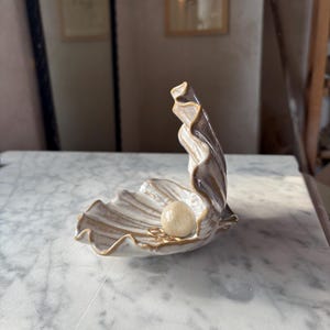 Może przedstawiać: Ceramiczna miseczka w kształcie muszli z błyszczącym białym wykończeniem i złotymi krawędziami. Naczynie posiada rzeźbioną, pionową stronę oraz płytką podstawę, w której znajduje się gładka, kremowa kula i mały złoty pierścionek.