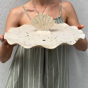 Könnte beinhalten: Handgefertigte, cremefarbene Keramik-Servierplatte mit Muscheldesign. Die Platte hat einen gewellten Rand und ist mit dunkelbraunen Punkten besprenkelt. Die Muschel befindet sich in der Mitte der Platte.