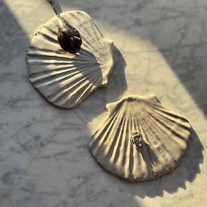 Può includere: Due piatti in ceramica bianco sporco a forma di conchiglia. Un piatto contiene un cucchiaio d'argento con dettagli ornati. L'altro piatto contiene un piccolo anello d'argento con un design decorativo. I piatti sono su una superficie di marmo.