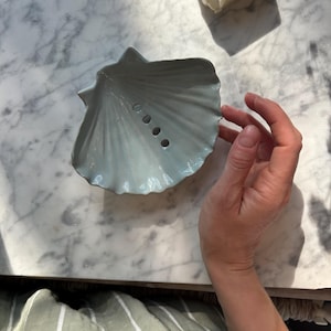 Può includere: Un portasapone a forma di conchiglia azzurra con tre fori di drenaggio. Il portasapone è in ceramica e ha una superficie strutturata. Una saponetta è visibile sullo sfondo. Progettato per contenere sapone e mantenerlo asciutto.