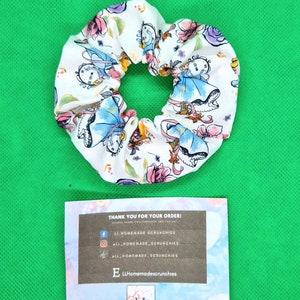 May include: White fabric scrunchie with a colorful print of Alice in Wonderland characters. The scrunchie is tied in a knot and has a small tag attached with the text "LL Homemadescrunchies".