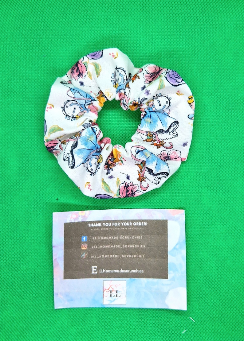 May include: White fabric scrunchie with a colorful print of Alice in Wonderland characters. The scrunchie is tied in a knot and has a small tag attached with the text "LL Homemadescrunchies".